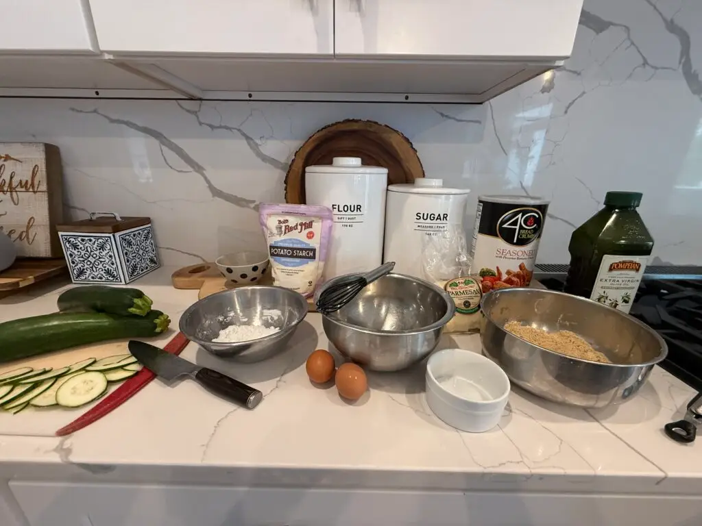 Dredging station set-up for frying zucchini on white quartz counter with backsplash, featuring potato starch, eggs, zucchini, breadcrumbs