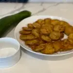 Plate of crispy fried zucchini with fresh whole zucchini and flakey salt in ceramic ramekin