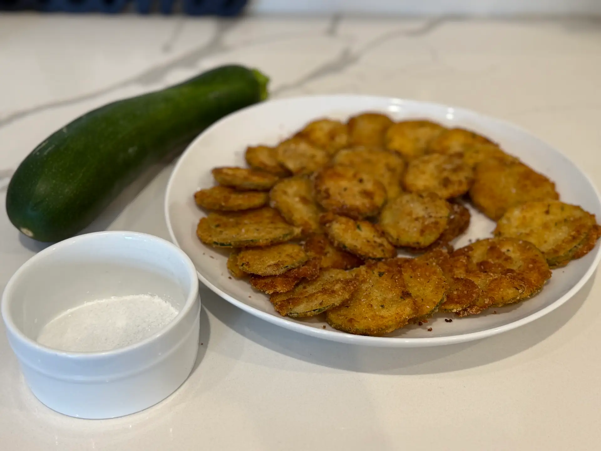 Plate of crispy fried zucchini with fresh whole zucchini and flakey salt in ceramic ramekin