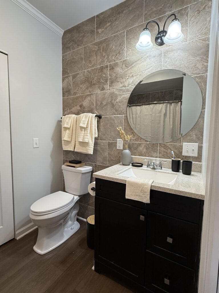Neutral, masculine bathroom, round mirror above dark wood vanity with light granite countertop, light towels and dark accent decor.
