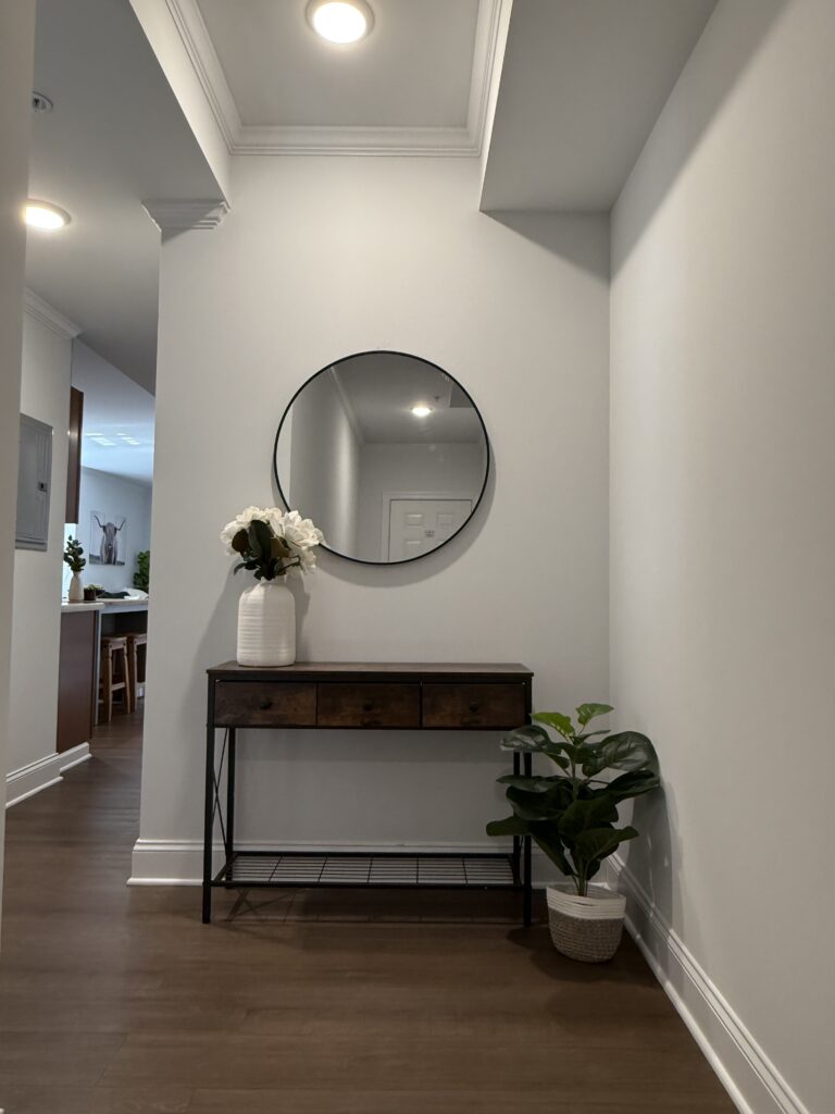 Entryway with round mounted mirror over wooden three drawer entry table with white vase and hydrangeas; fiddlehead fern in canvas pot beside table on the floor