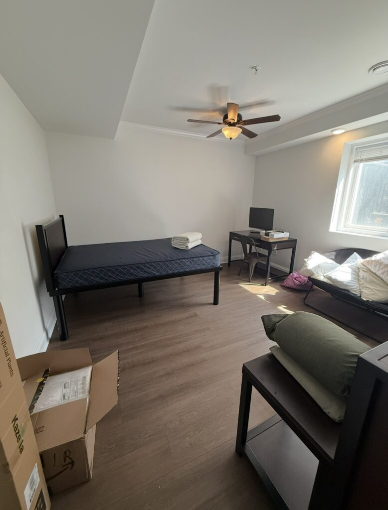 Unfinished bedroom with mattress on bed, furniture, but no decor, no rug, laminate wood floors; ceiling fan with light and large window