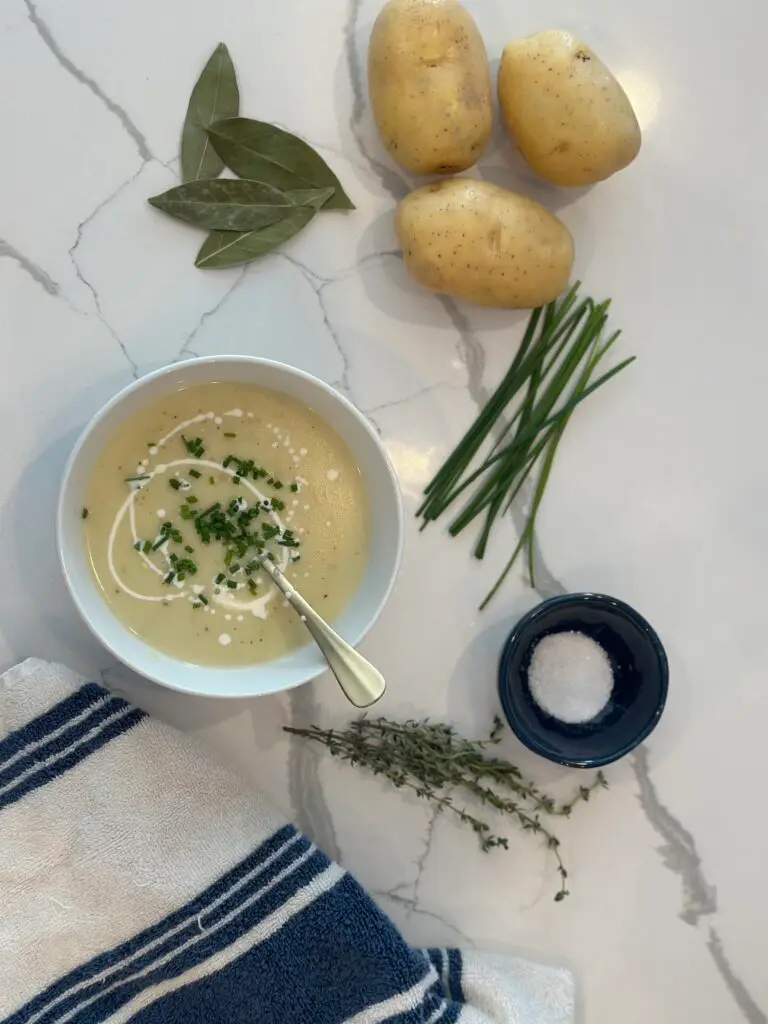 potato leek soup styled with fresh potatoes, chives, fresh herbs, salt and towel on white quartz counter