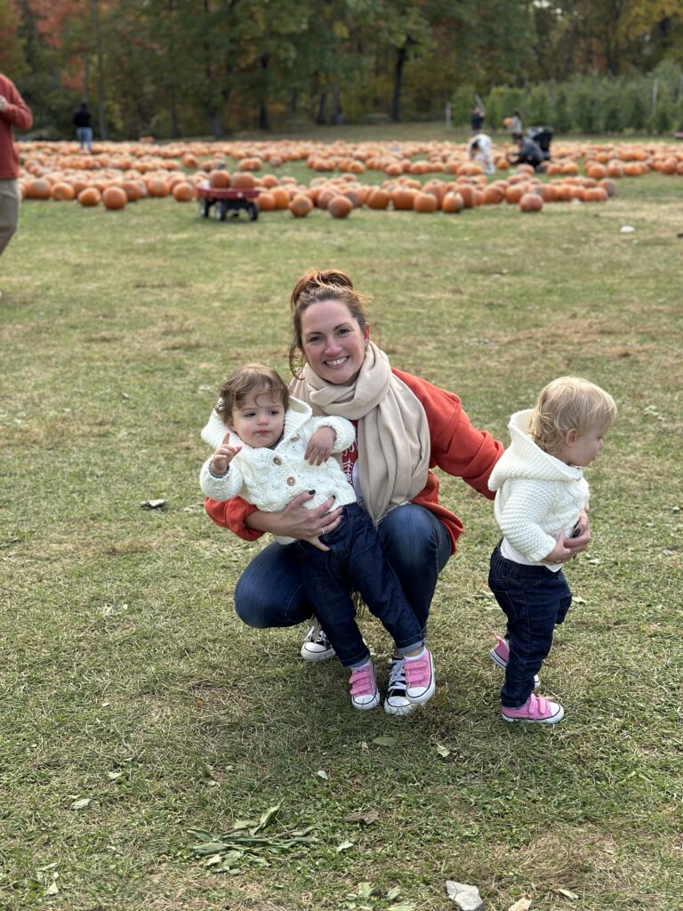 Mom in fall colors in pumpkin field, wrangling twin girls in cream sweaters for a picture