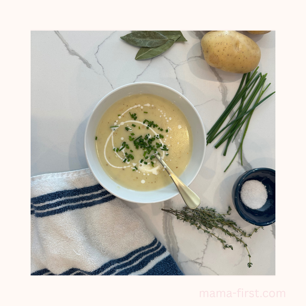 vichyssoise soup in white bowl on white quartz counter with potatoes, chives, thyme, salt, bay leaf, & all clad white & blue towel surrounding