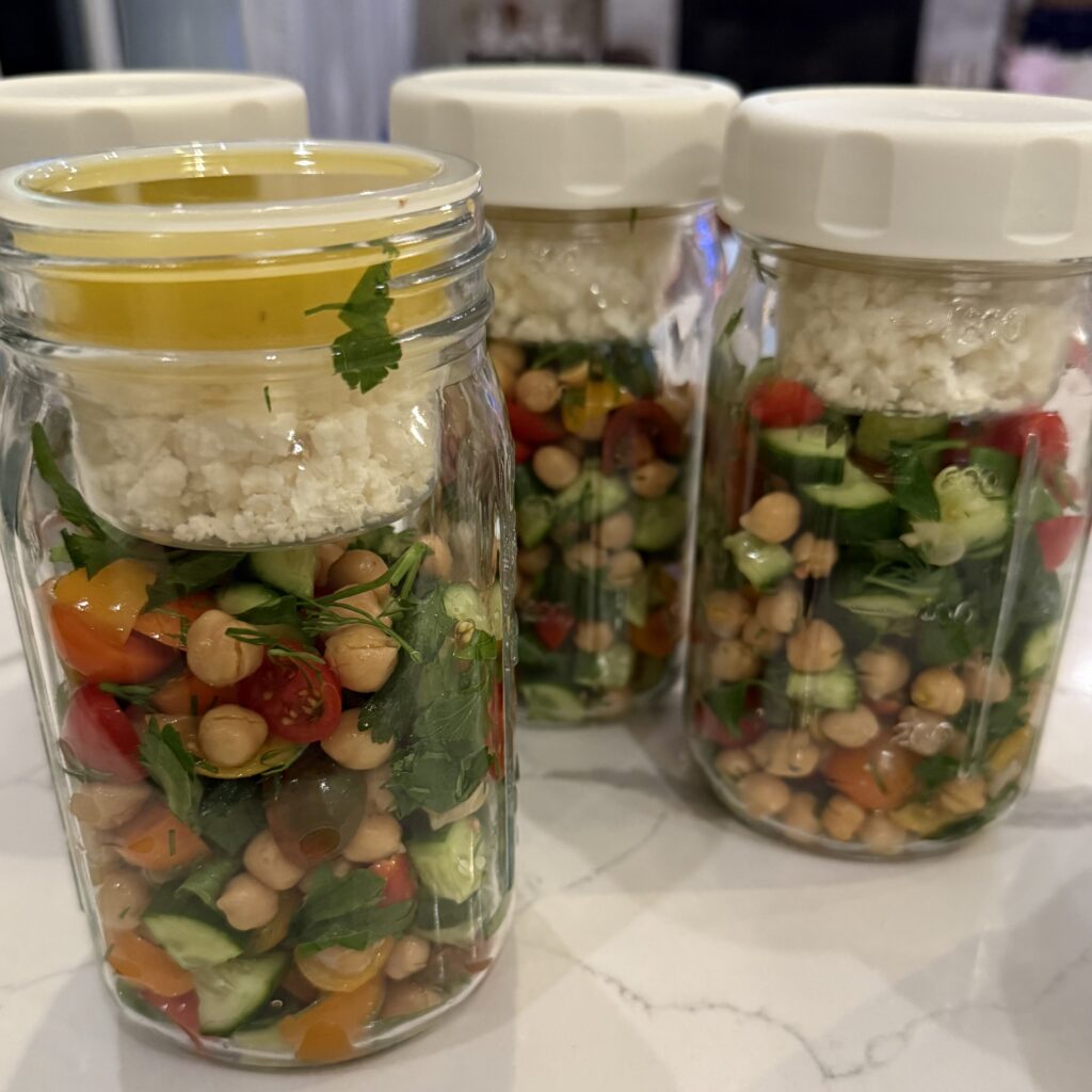 Layered mason jar salads with chickpeas, cucumber, tomatoes and herbs.  feta and dressing separated with extra compartments.