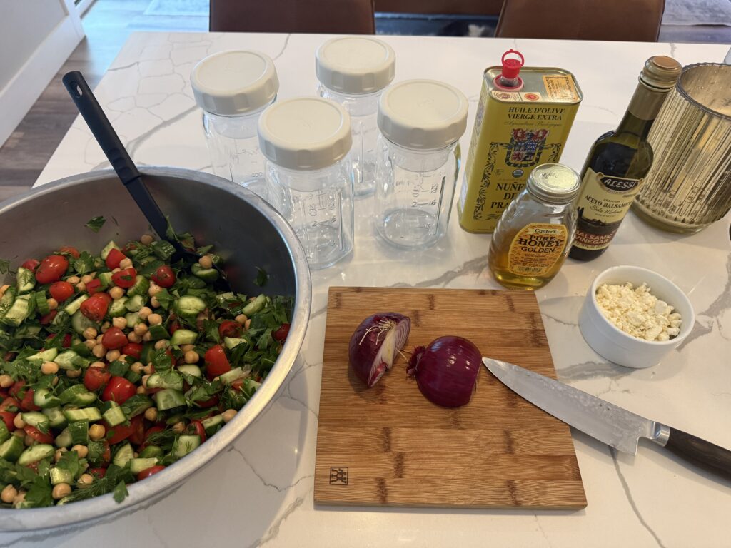 Salad lunch prep set up with Mediterranean Salad in bowl, cutting board with red onion and knife, olive oil, balsamic vinegar & honey for dressing, Glass mason jars for meal prep storage