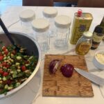 Salad lunch prep set up with Mediterranean Salad in bowl, cutting board with red onion and knife, olive oil, balsamic vinegar & honey for dressing, Glass mason jars for meal prep storage