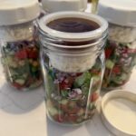 Glass Mason jar filled with chickpea, cucumber, tomato and herb salad. Insert container separate the feta cheese crumbles and dressing. Cream lid keeps everything contained.