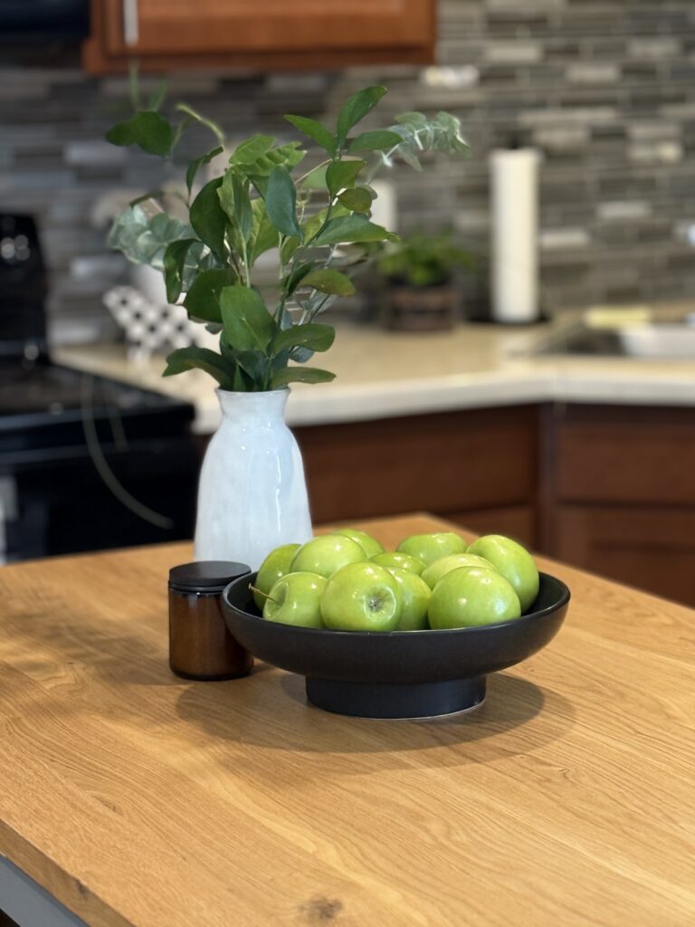 portrait shot of a grouping of a vase with greenery, a black pedestal bowl filled with green apples and a candle, all on a light wood center island 