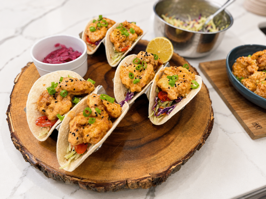 Crispy bang bang shrimp street tacos with creamy Thai chili sauce, broccoli slaw, scallions, and black sesame seeds served on a rustic wooden platter.