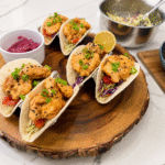 Crispy bang bang shrimp street tacos with creamy Thai chili sauce, broccoli slaw, scallions, and black sesame seeds served on a rustic wooden platter.