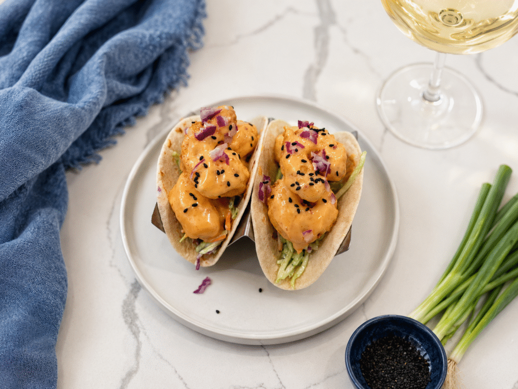 Crispy bang bang shrimp street tacos with creamy Thai chili sauce, broccoli slaw, black sesame seeds, and red onion, styled with white wine, fresh scallions, and a blue linen napkin.