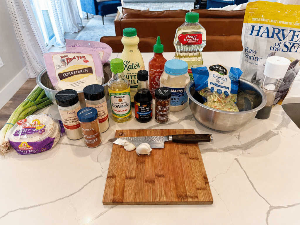 Ingredients Shot. From left to right: fresh scallions, mission street flour tortillas, cornstarch, onion powder, paprika, garlic powder, rice vinegar, buttermilk, fish sauce, sriracha, red chili flakes, Hellmann's Light mayonnaise, canola oil, Broccoli slaw, Harvest of the Sea frozen raw shrimp, salt and pepper and in front a bamboo cutting board with a knife and four cloves of garlic ready to be chopped.