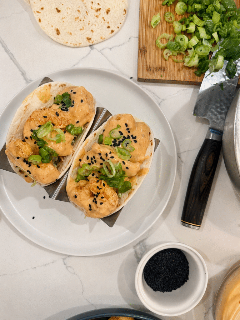 Prepared shrimp tacos topped with black sesame & scallion in stainless holder on white plate. Styled with knife, cutting board with chopped scallions, soft tortillas fanned and black sesame in ramekin. 
