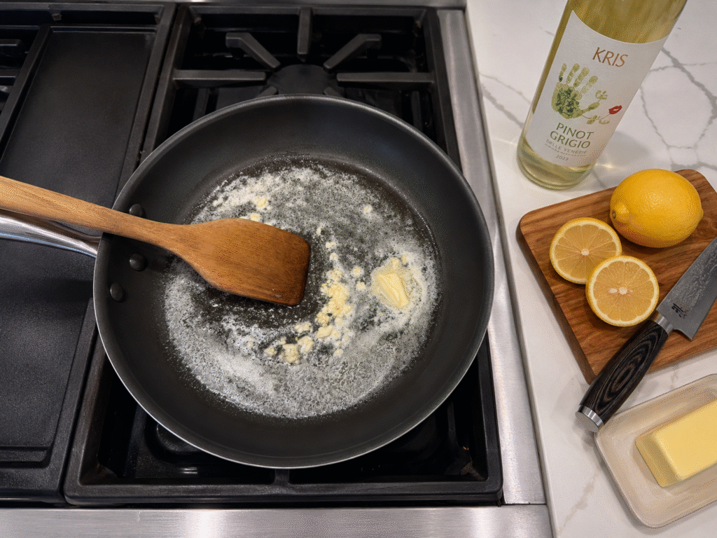 Garlic sautéing in butter in a skillet for homemade lemon butter sauce, served with stuffed monkfish. Fresh lemons, butter, and Pinot Grigio are arranged beside the stove during recipe preparation.