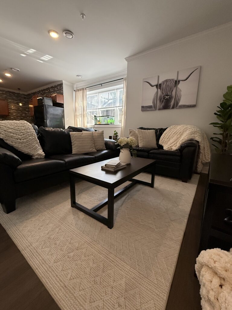 After shot 2: Living room over looking into kitchen.  Farmhouse vibe living room; light cream throw pillows on couches, chunky knit throws draped on couches and out of wicker basket on floor, cute vase with flowers and book boxes on table, horned steer wall art and greenery completing the space