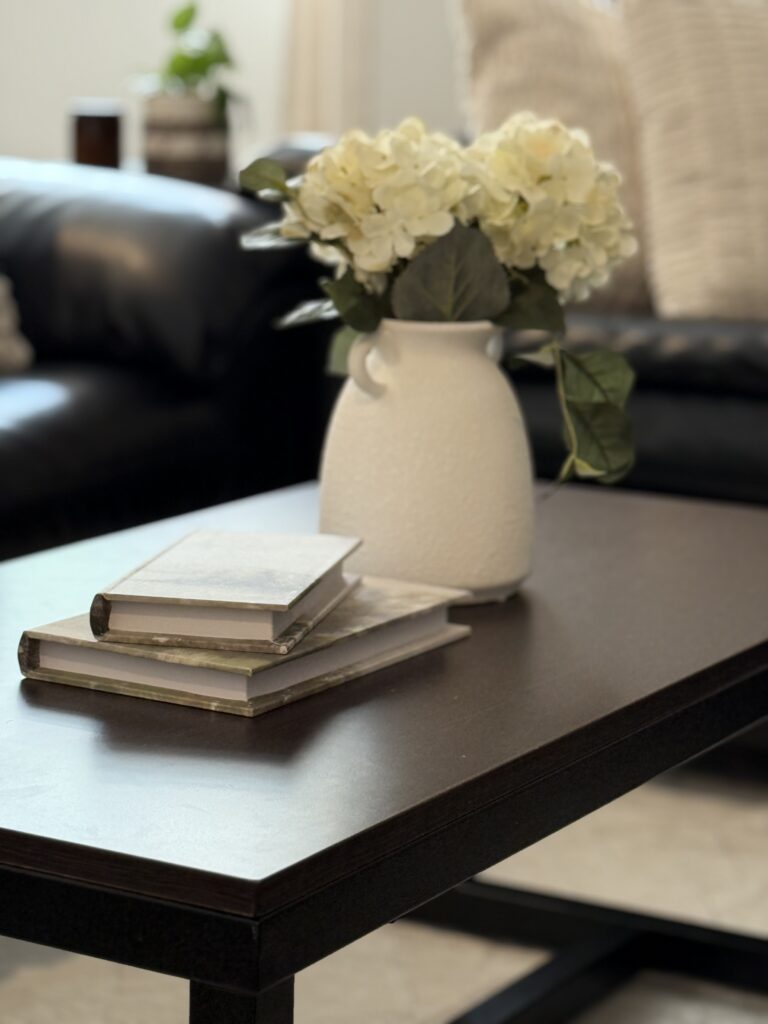 coffee table close up with hydrangea filled vase, decorative books, throw pillows and plants faded in background on and next to couch