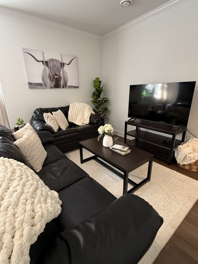 After shot: Farmhouse vibe living room; light cream throw pillows on couches, chunky knit throws draped on couches and out of wicker basket on floor, cute vase with flowers and book boxes on table, horned steer wall art and greenery completing the space