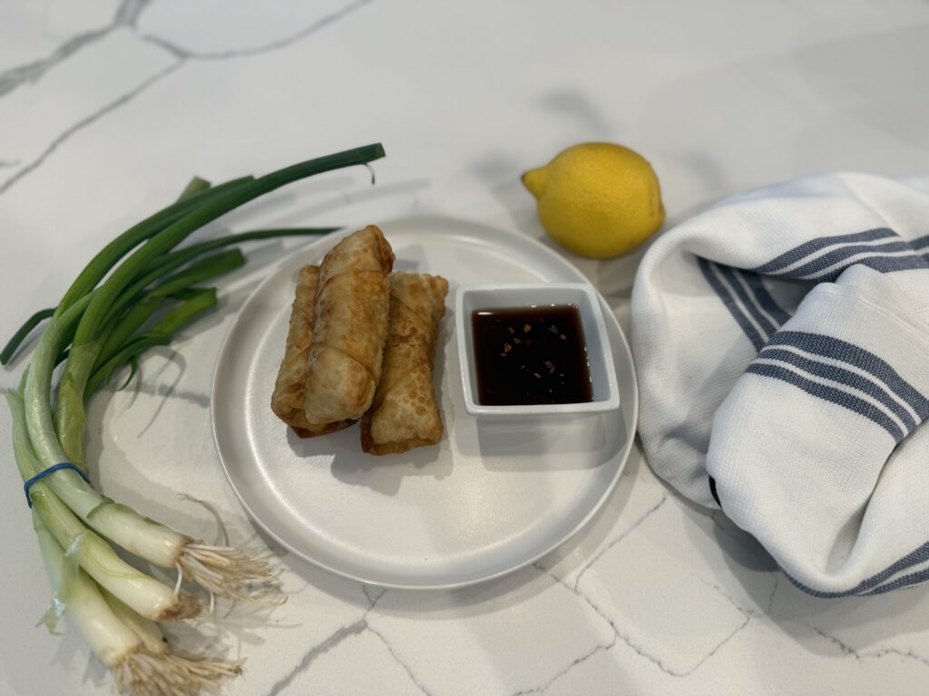 Three crispy fried crab egg rolls stacked on white plate, next to shallow white ramekin of sweet & spicy cherry dipping sauce. Styled with scallions bunch to left of plate, lemon at 1 o'clock and white towel with blue striping to the right.