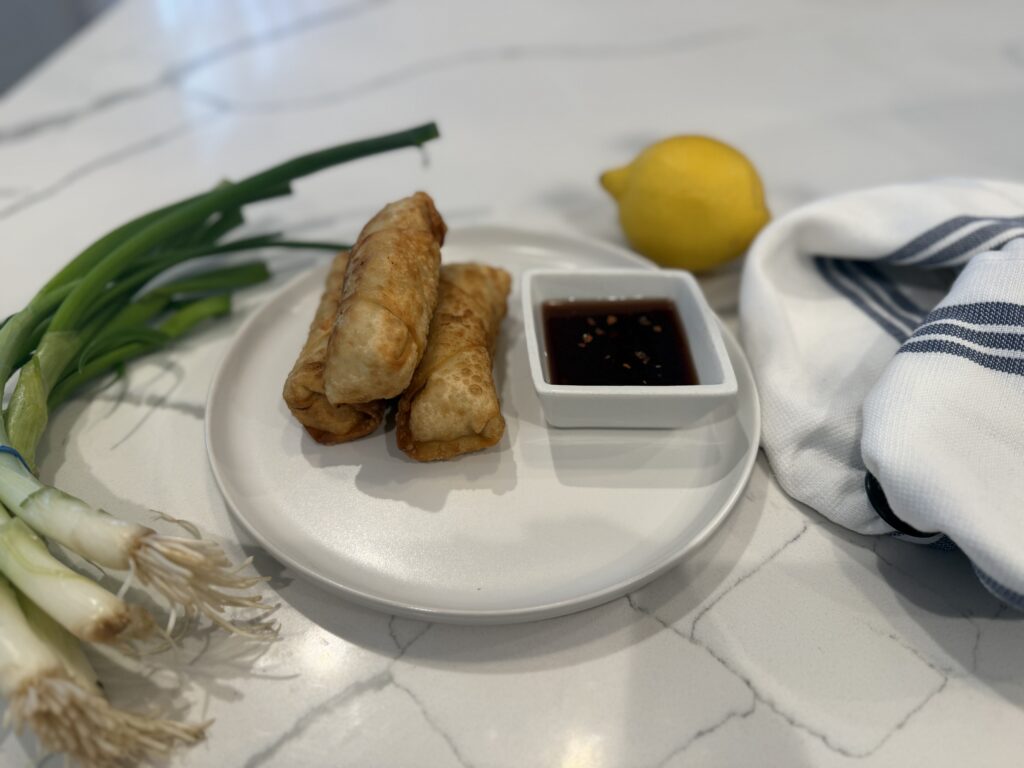 Crab Rangoon Rolls artfully styled on white plate with white ramekin filled with spicy cherry dipping sauce with scallions, lemon and towel placed around plate. 