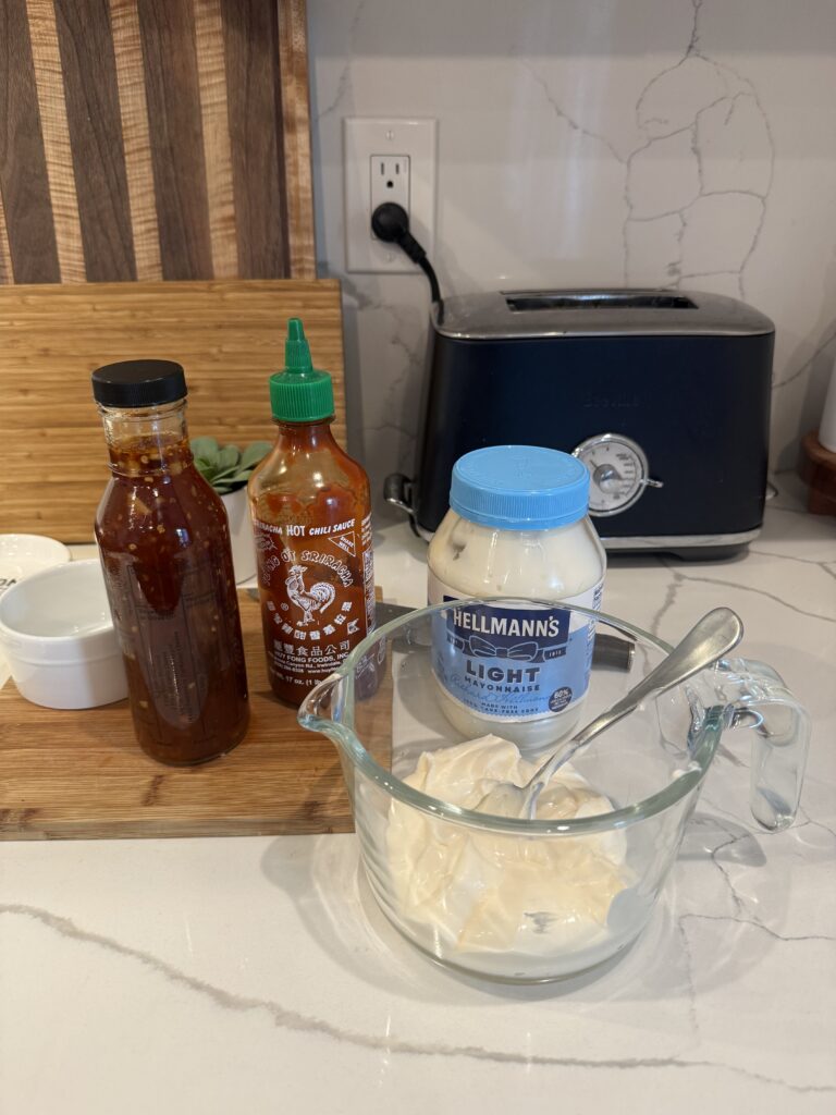 Bang Bang Sauce set-up: Bottled homemade Thai chili sauce, Sriracha, Mayo jar, and glass measuring cup filled with mayo on white counter with wooden cutting boards. 