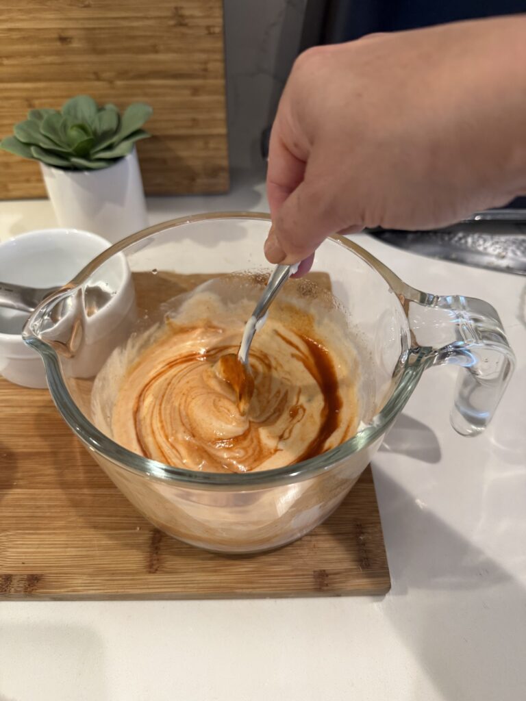Sriracha creating a pretty swirl while being mixed into mayo in glass measuring cup on wooden cutting board