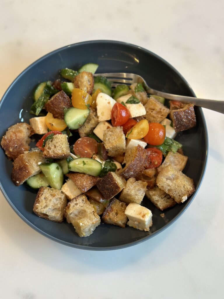 Blue single serving ceramic bowl of panzanella salad: toasted sourdough, fresh mozzarella, tomatoes, cucumbers, and basil.