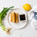 Crispy Crab "Rangoon" Rolls styled on white ceramic plate with white ramekin of cherry dipping sauce, artfully surrounded by a scallion bunch, fresh lemon, and white and blue chef's towel.